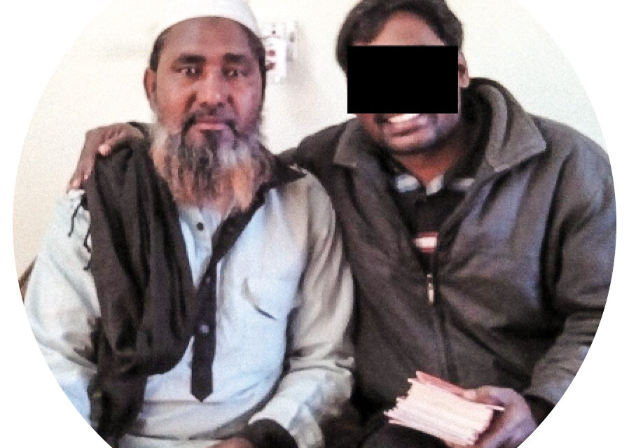 Two men sitting together smiling and one has a black box over his eyes and is holding a bible
